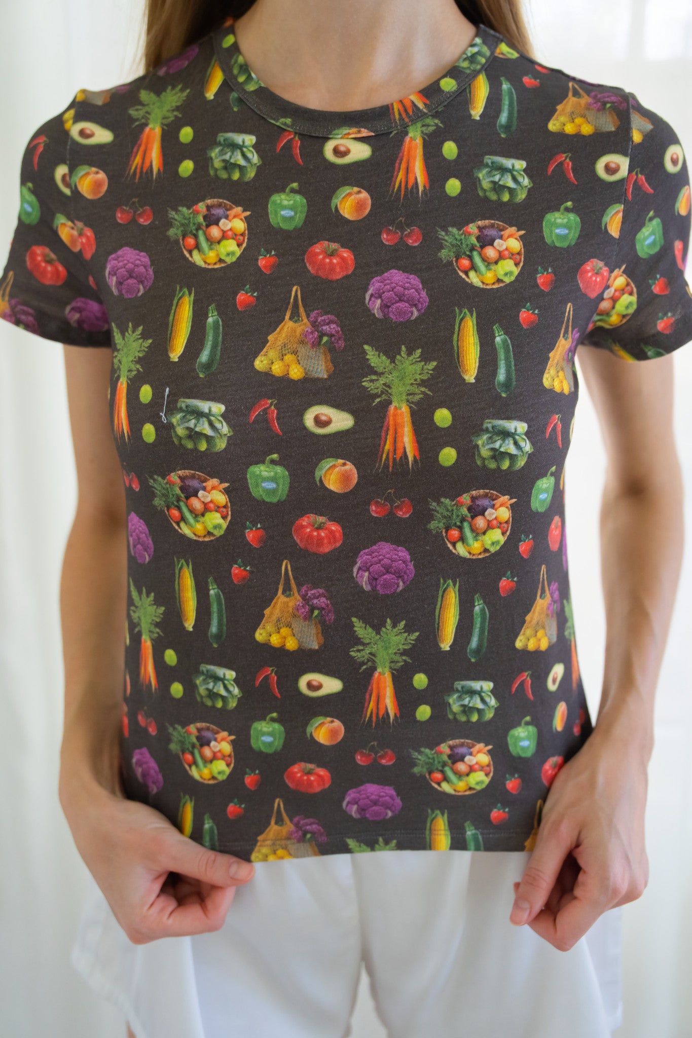 A person wearing a dark charcoal gray t-shirt with a colorful all-over print of various fruits and vegetables, paired with white shorts.