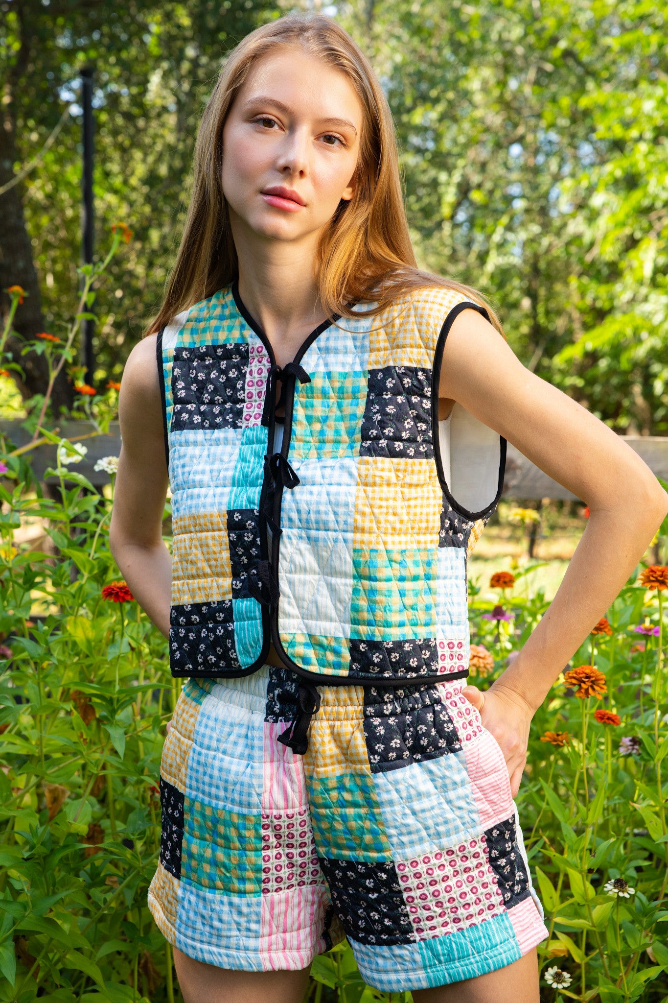 Person standing outdoors wearing a sleeveless patchwork quilted vest and matching patchwork quilted shorts. The outfit features a colorful grid pattern of various squares in blue, yellow, pink, black, and white, with black trim and a black tie at the collar.