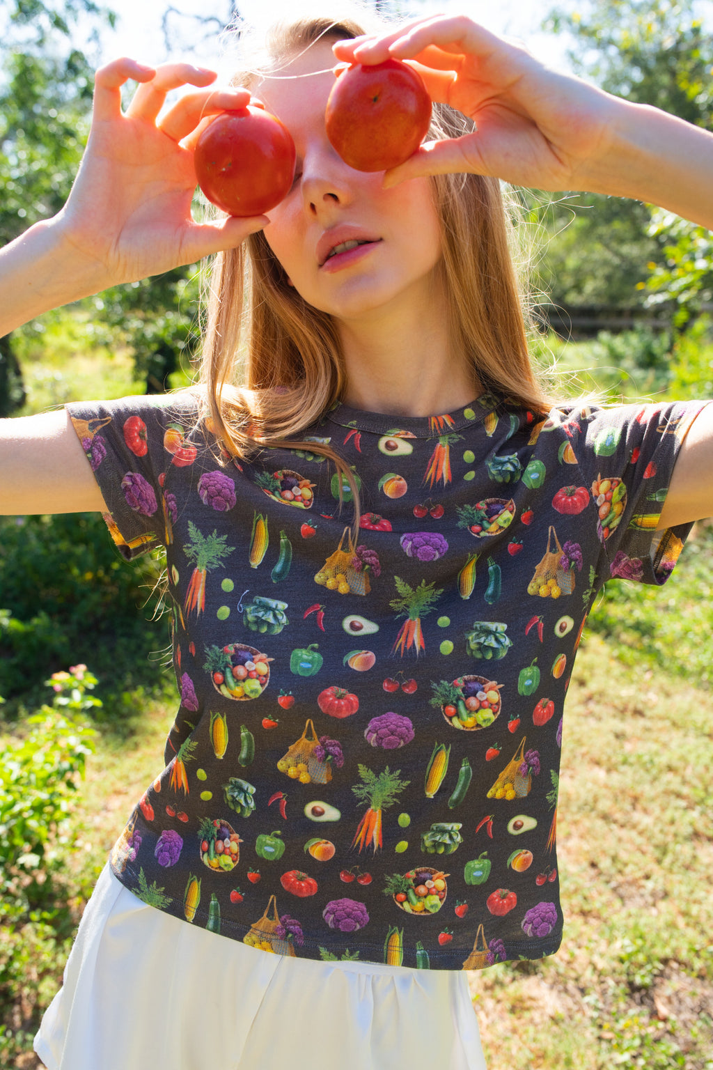 A woman outdoors wearing a dark gray t-shirt with a colorful all-over print of vegetables, holding two red tomatoes to her eyes like glasses.