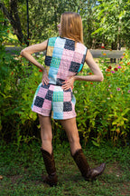 Person standing outdoors in a garden wearing a sleeveless patchwork quilted vest and matching patchwork quilted shorts, paired with brown cowboy boots.
