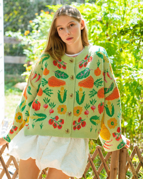 Person wearing a light green knit cardigan with a colorful vegetable and floral pattern, paired with a white ruffled skirt.