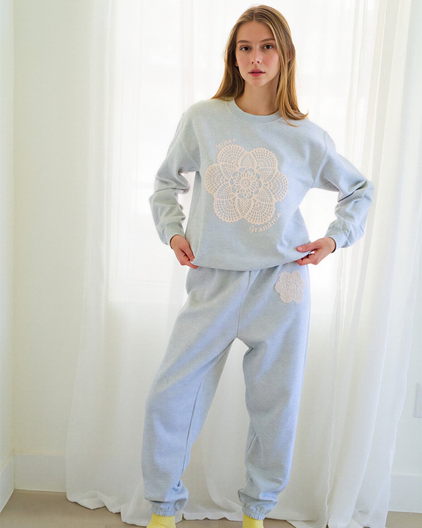 Person wearing a light grey sweatshirt and sweatpants set with a large cream crochet floral design on the chest and a matching pocket detail on the thigh, paired with yellow socks, standing in front of white curtains.