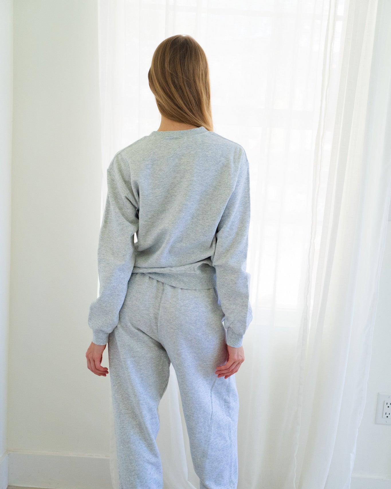 Back of person wearing a light heather gray sweatshirt and matching jogger sweatpants, standing barefoot in yellow slippers in a bright room with white curtains.