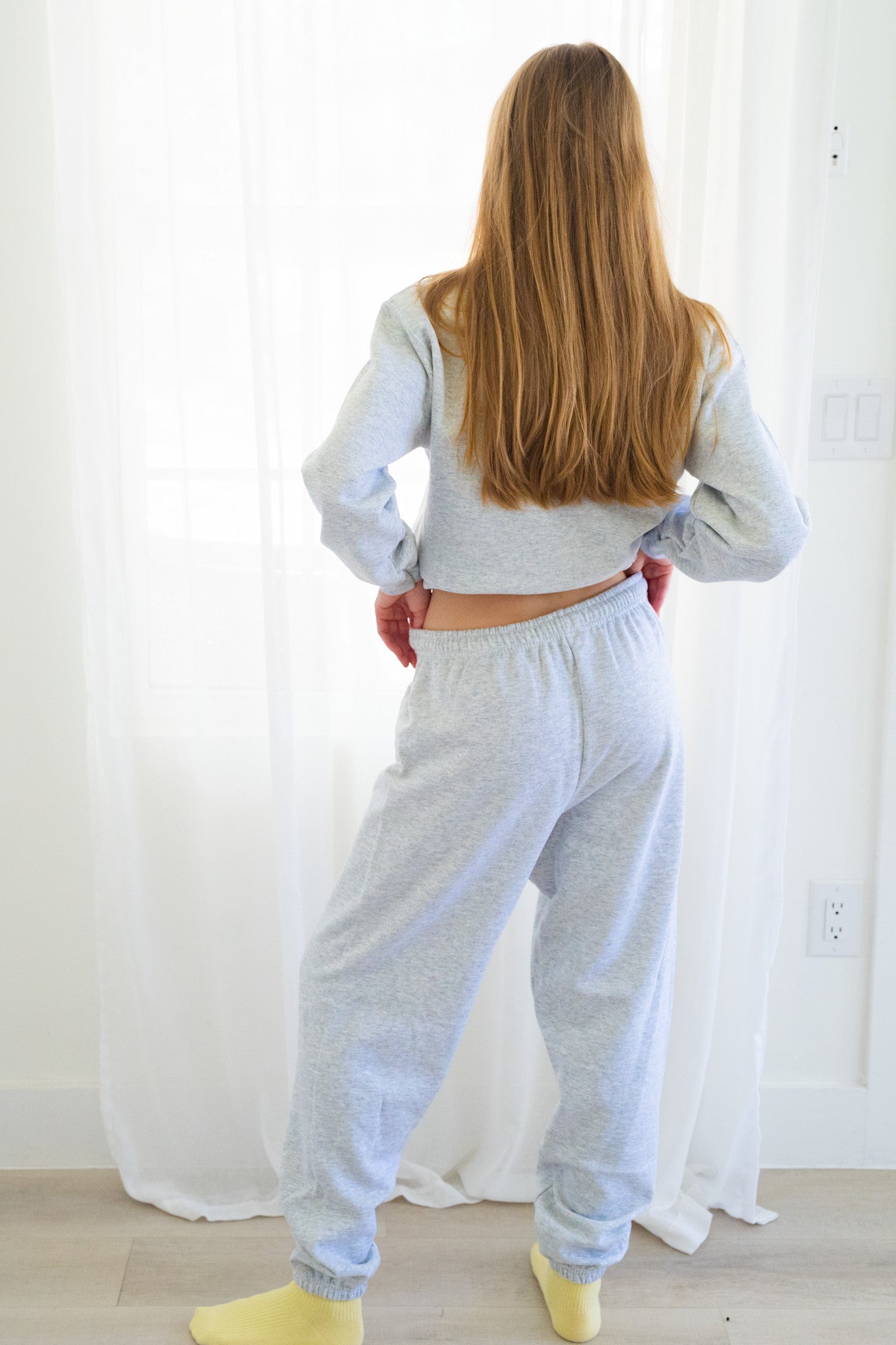 Back of person wearing a light gray heather sweatshirt and matching sweatpants, standing in front of white sheer curtains.