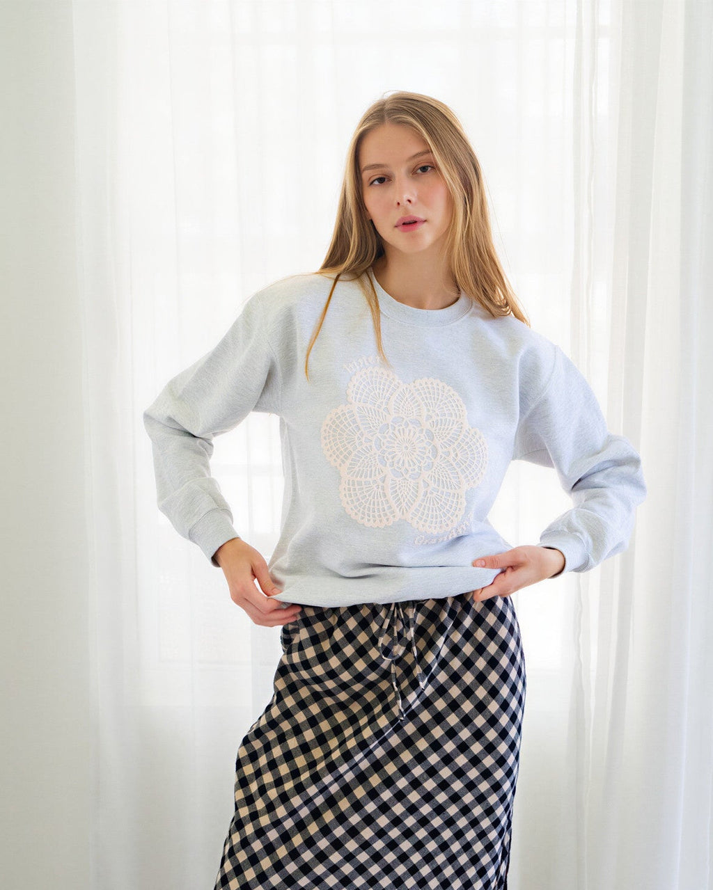 Person wearing a light gray crewneck sweatshirt with a white floral embroidery on the front, paired with a black and white gingham midi skirt and dark blue flats, standing in front of white curtains.