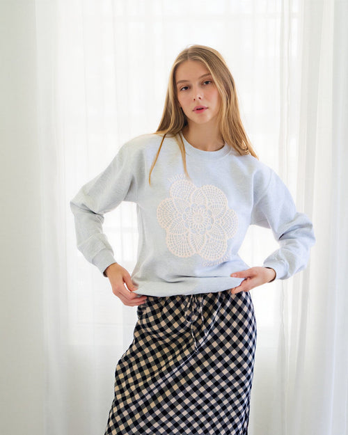 Person wearing a light gray crewneck sweatshirt with a white floral embroidery on the front, paired with a black and white gingham midi skirt and dark blue flats, standing in front of white curtains.