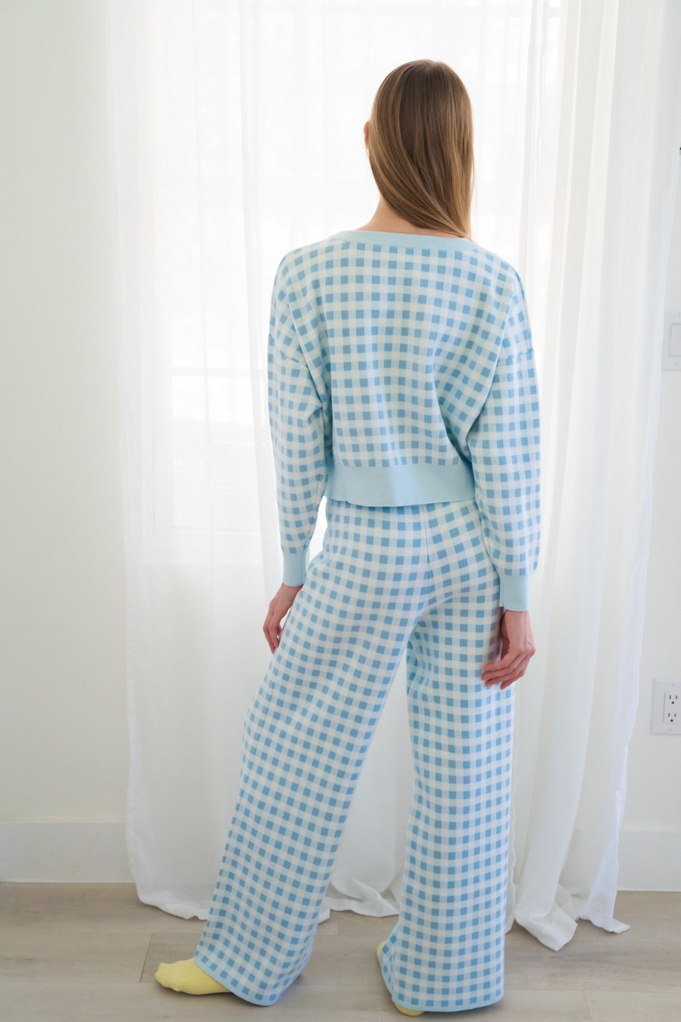 Back of person wearing a light blue and white gingham two-piece lounge set  in front of white sheer curtains.