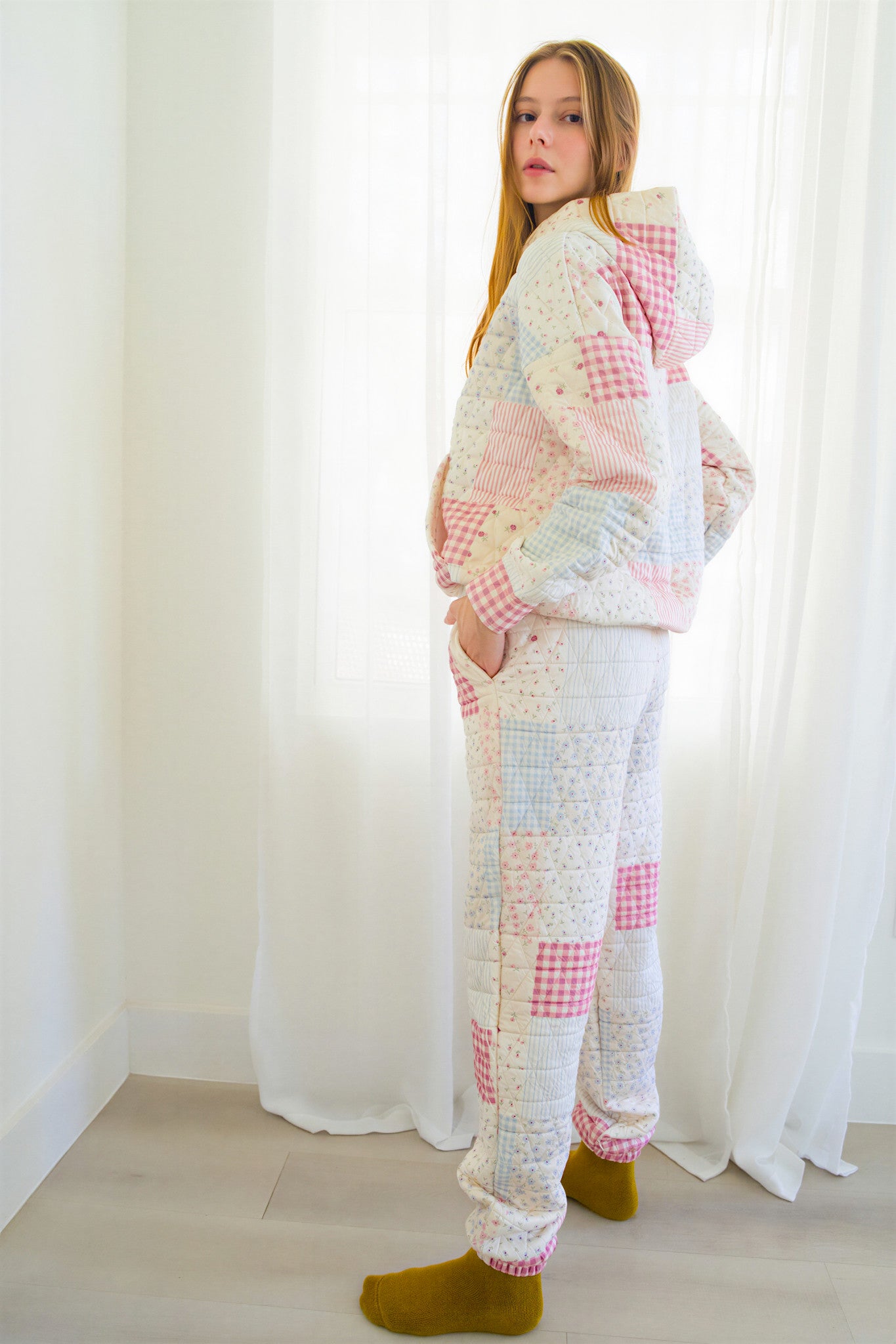 Person standing in a bright room wearing a pastel patchwork quilted hoodie and matching jogger pants with olive green socks.
