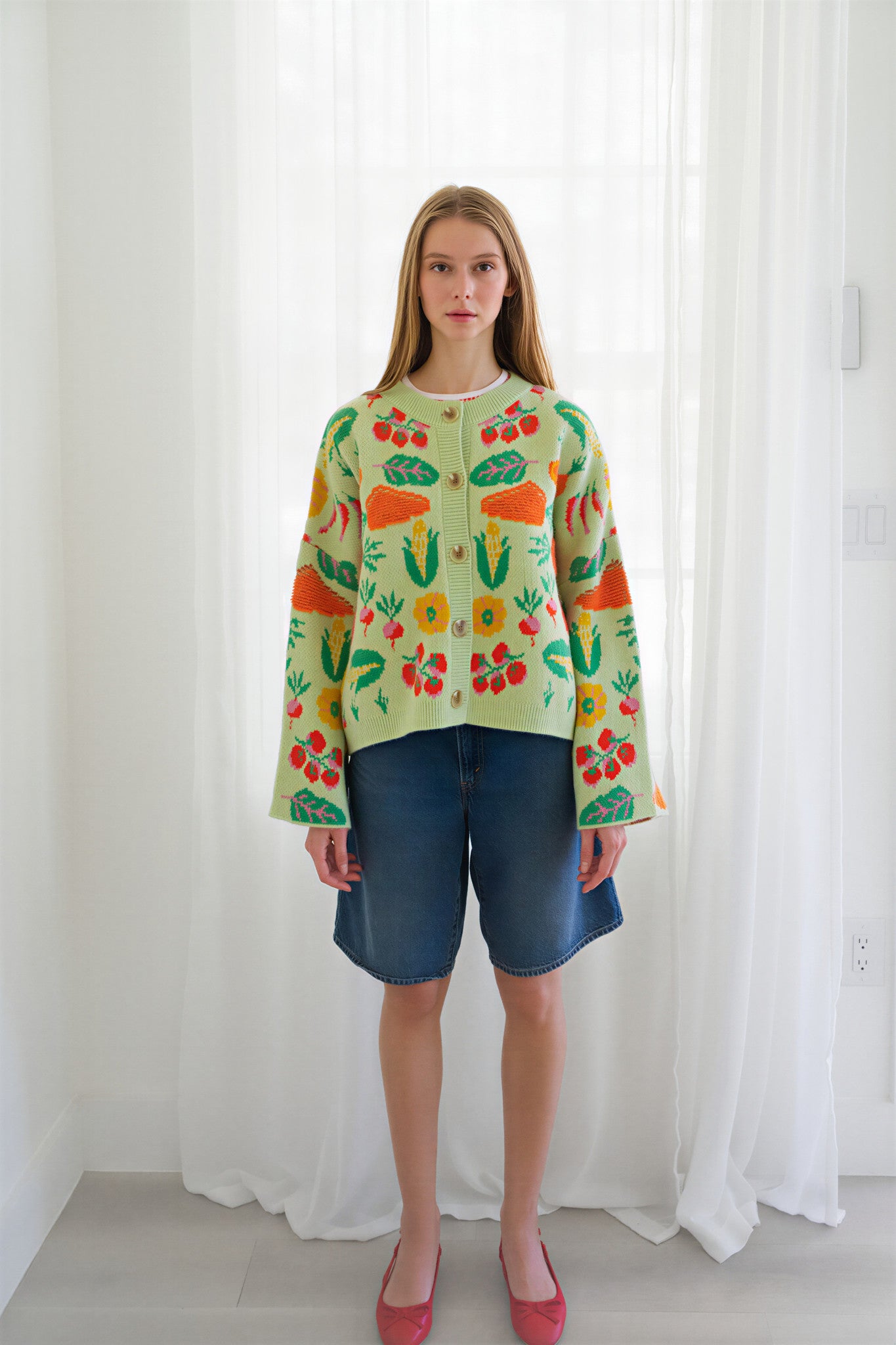 Person wearing a light green knit cardigan with a colorful vegetable pattern, paired with blue jeans and red shoes, standing in front of a white curtain background.