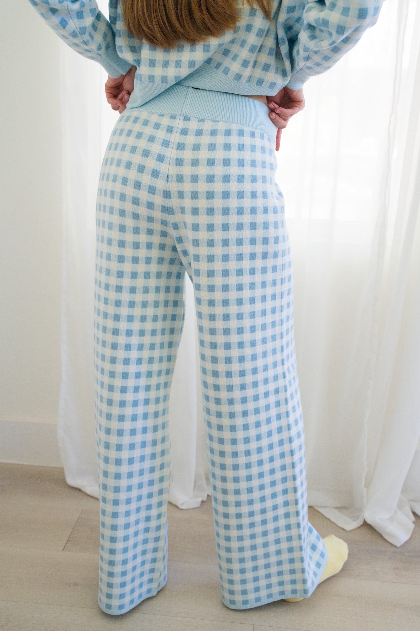 Back of person wearing a light blue and white gingham check pajama set with wide-leg pants standing in a bright room with white curtains and light wood flooring.