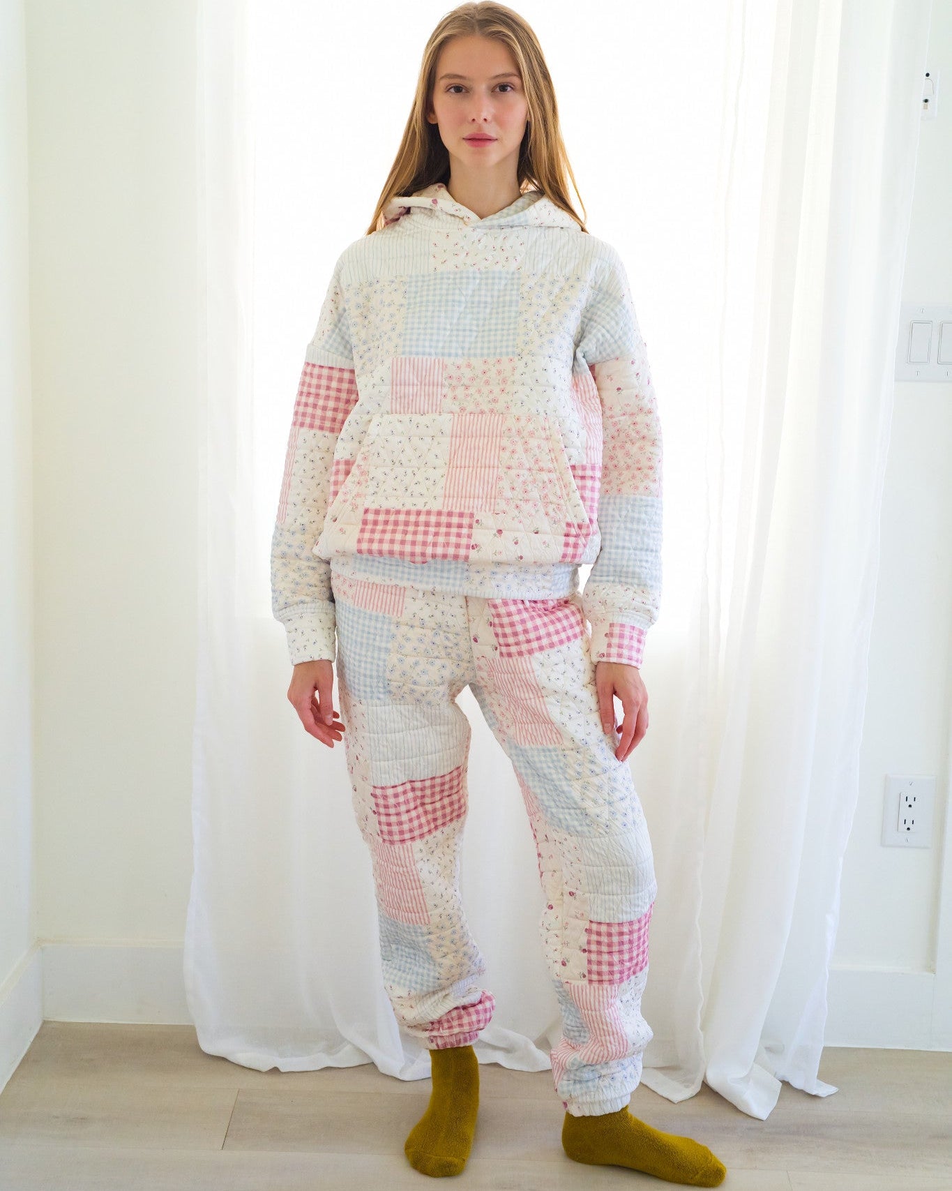 Person standing in a bright room wearing a matching patchwork hoodie and jogger pants set in pastel pink, white, and light blue fabrics, paired with olive green socks.