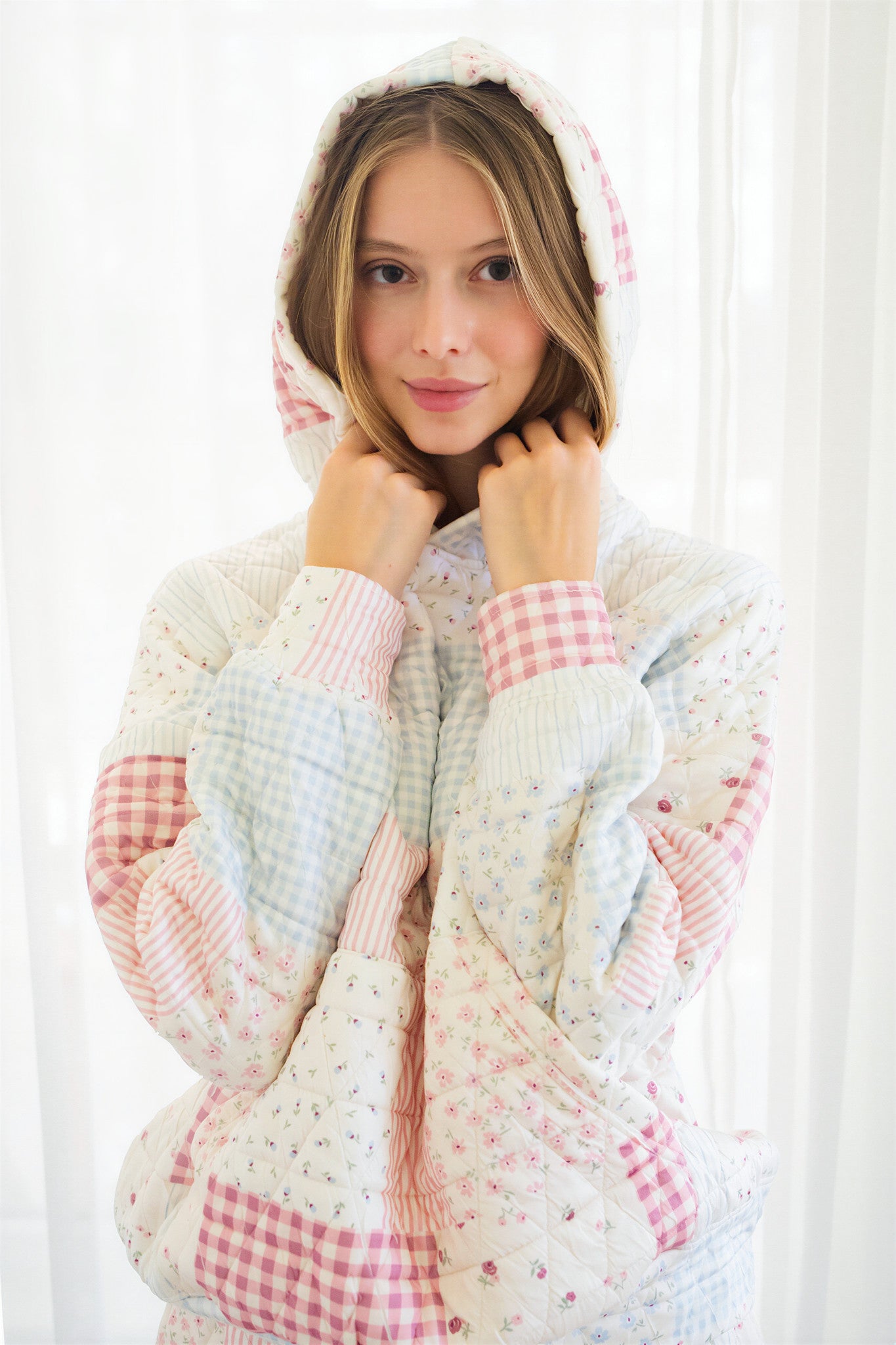 Person wearing a pastel patchwork quilted hooded jacket with pink, blue, and white gingham and floral patterns, posing in front of a white curtain.