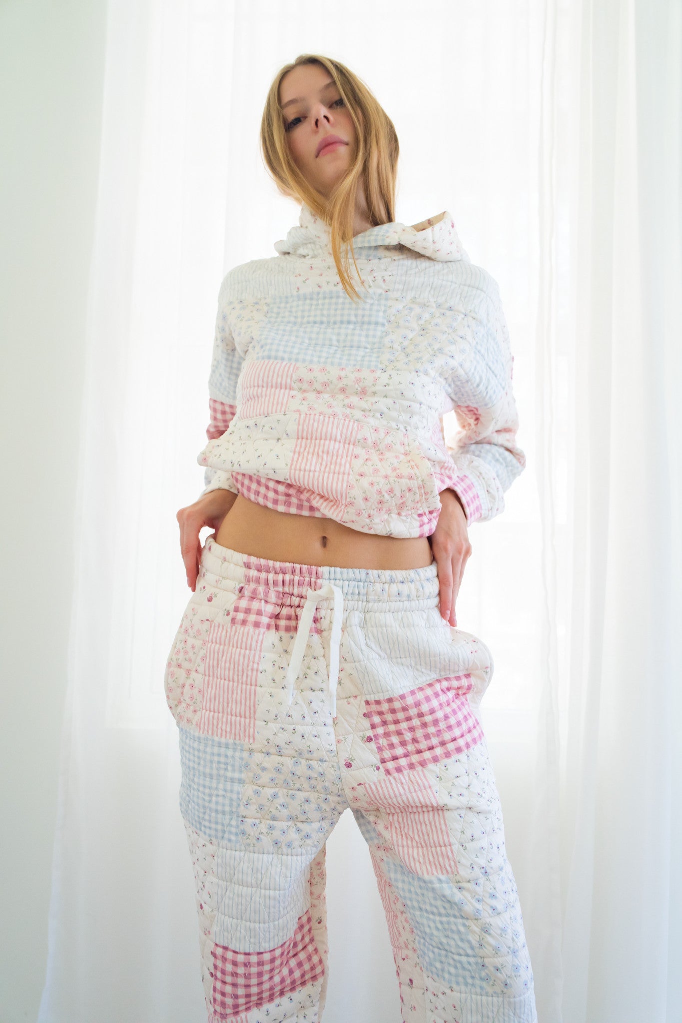 Person modeling a matching patchwork lounge set consisting of a cropped long-sleeve hoodie and drawstring pants in soft pastel colors including pink, blue, and white.