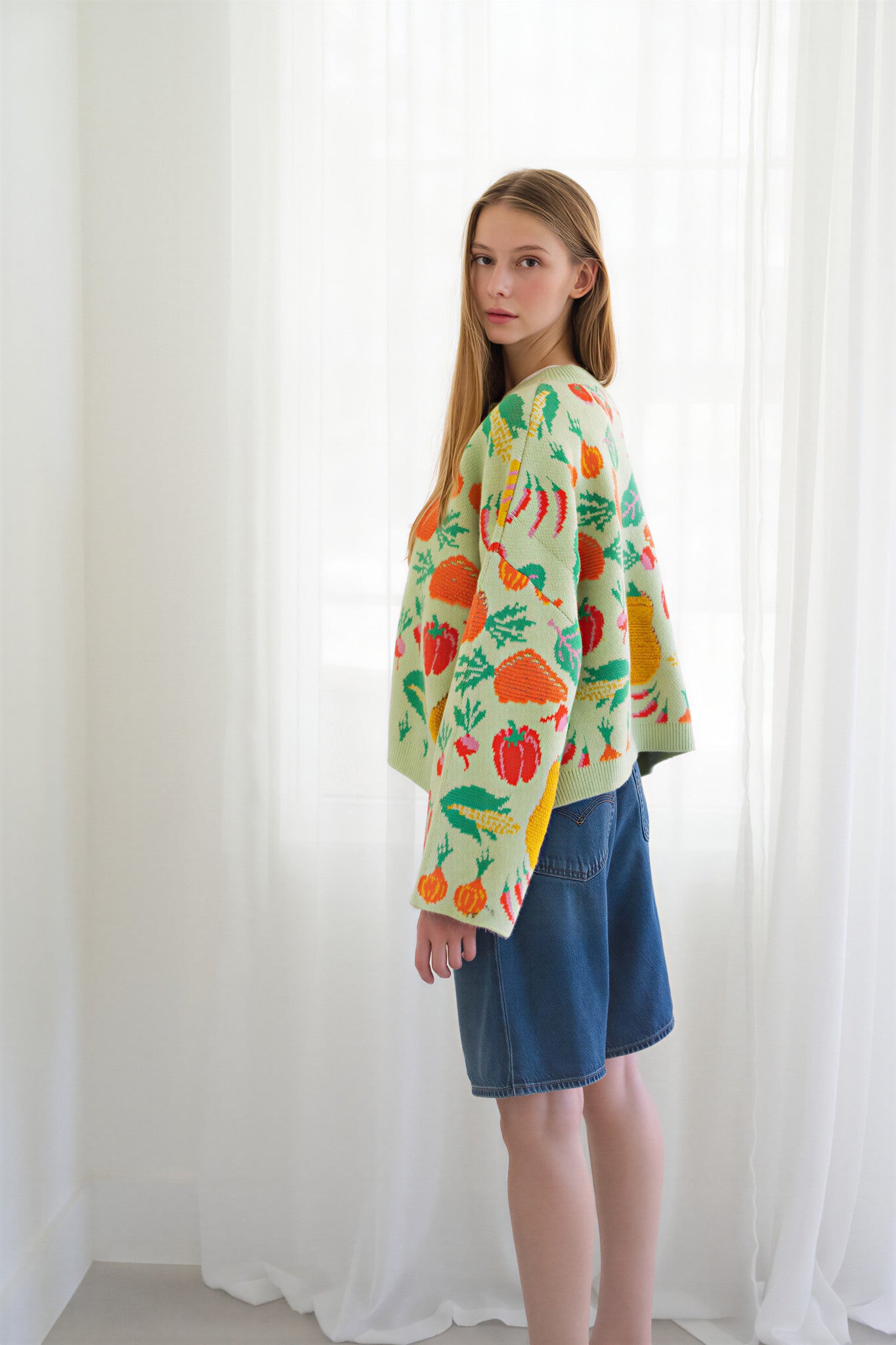 Side of person wearing a light green knit cardigan with a colorful vegetable pattern, paired with blue jeans, standing in front of a white curtain background.