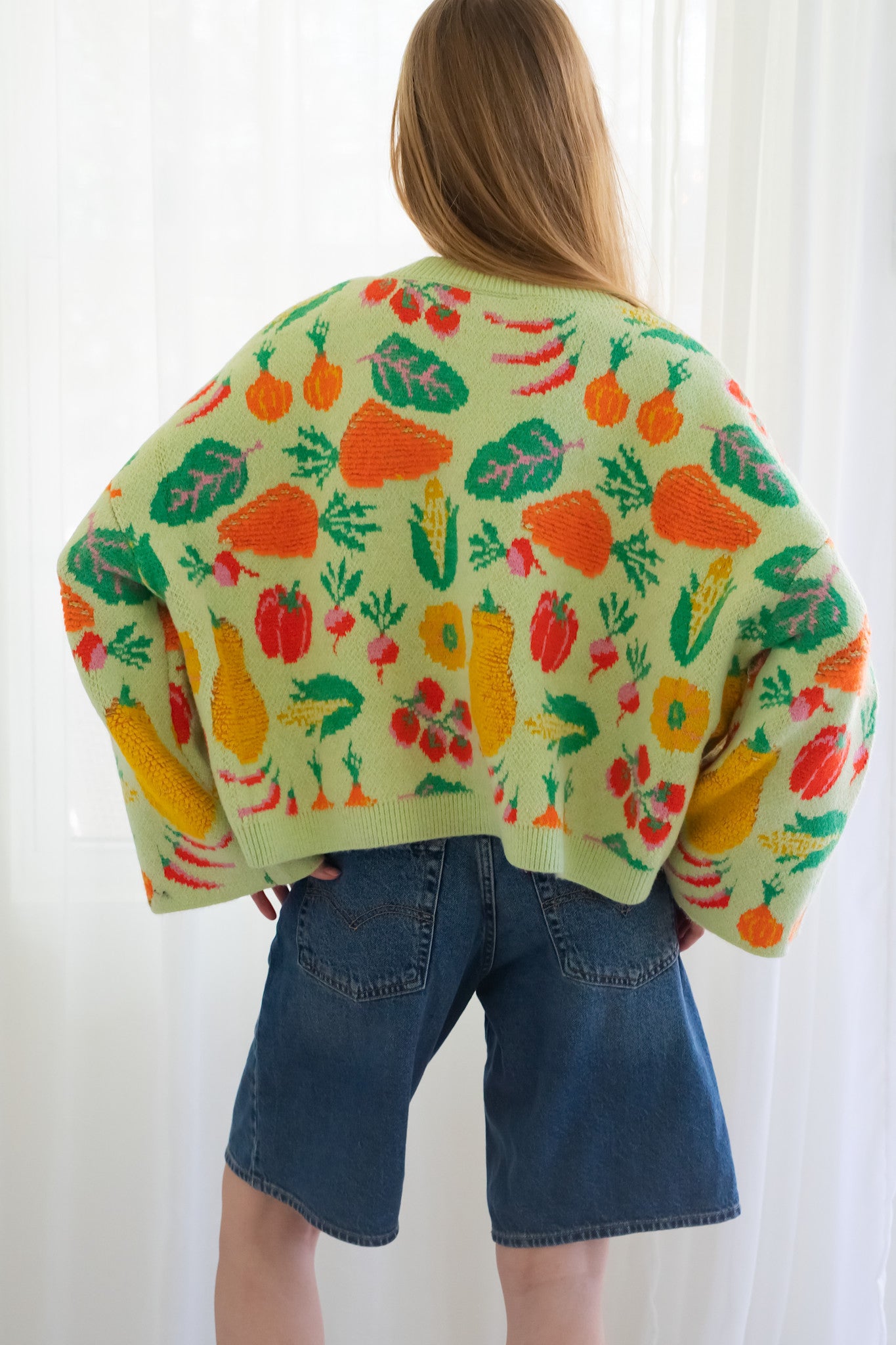 Back of person wearing a light green knit cardigan with a colorful vegetable pattern, paired with blue jeans, standing in front of a white curtain background.