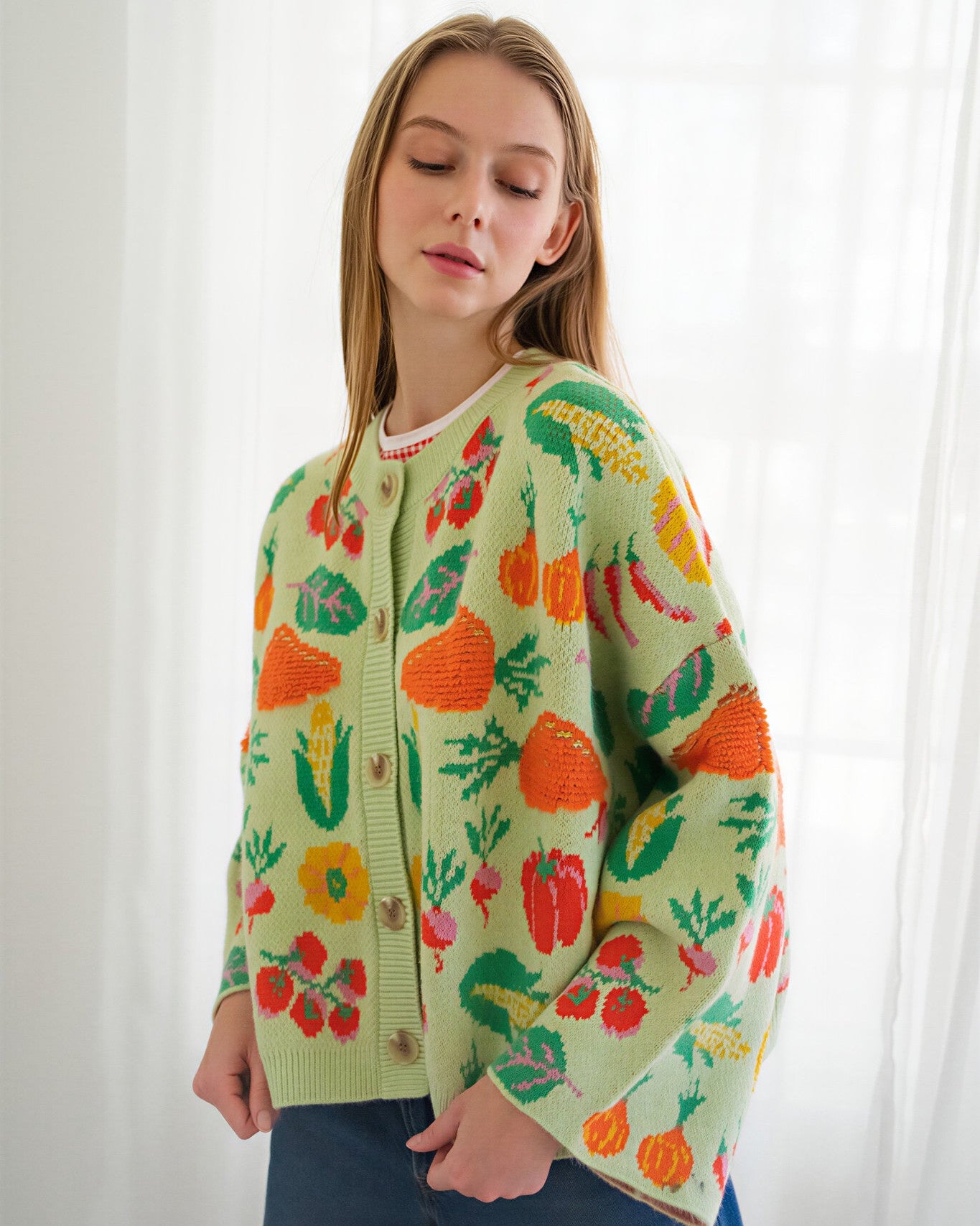 Person wearing a light green knit cardigan with a colorful vegetable pattern, paired with blue jeans, standing in front of a white curtain background.