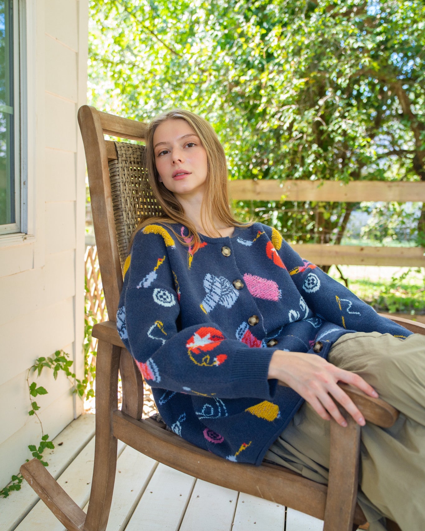 Person sitting in a wooden rocking chair on a porch, wearing a navy blue long-sleeve sweater with a colorful fishing-themed pattern and khaki pants.