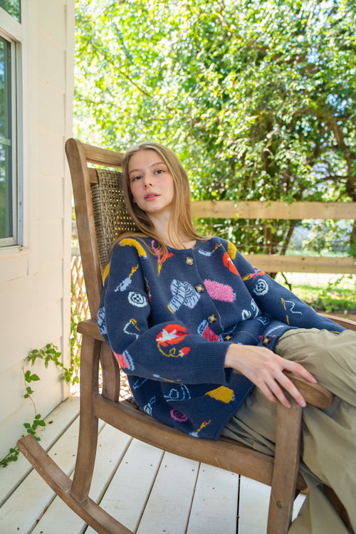 Person sitting in a wooden rocking chair on a porch, wearing a navy blue long-sleeve sweater with a colorful fishing-themed pattern and khaki pants.