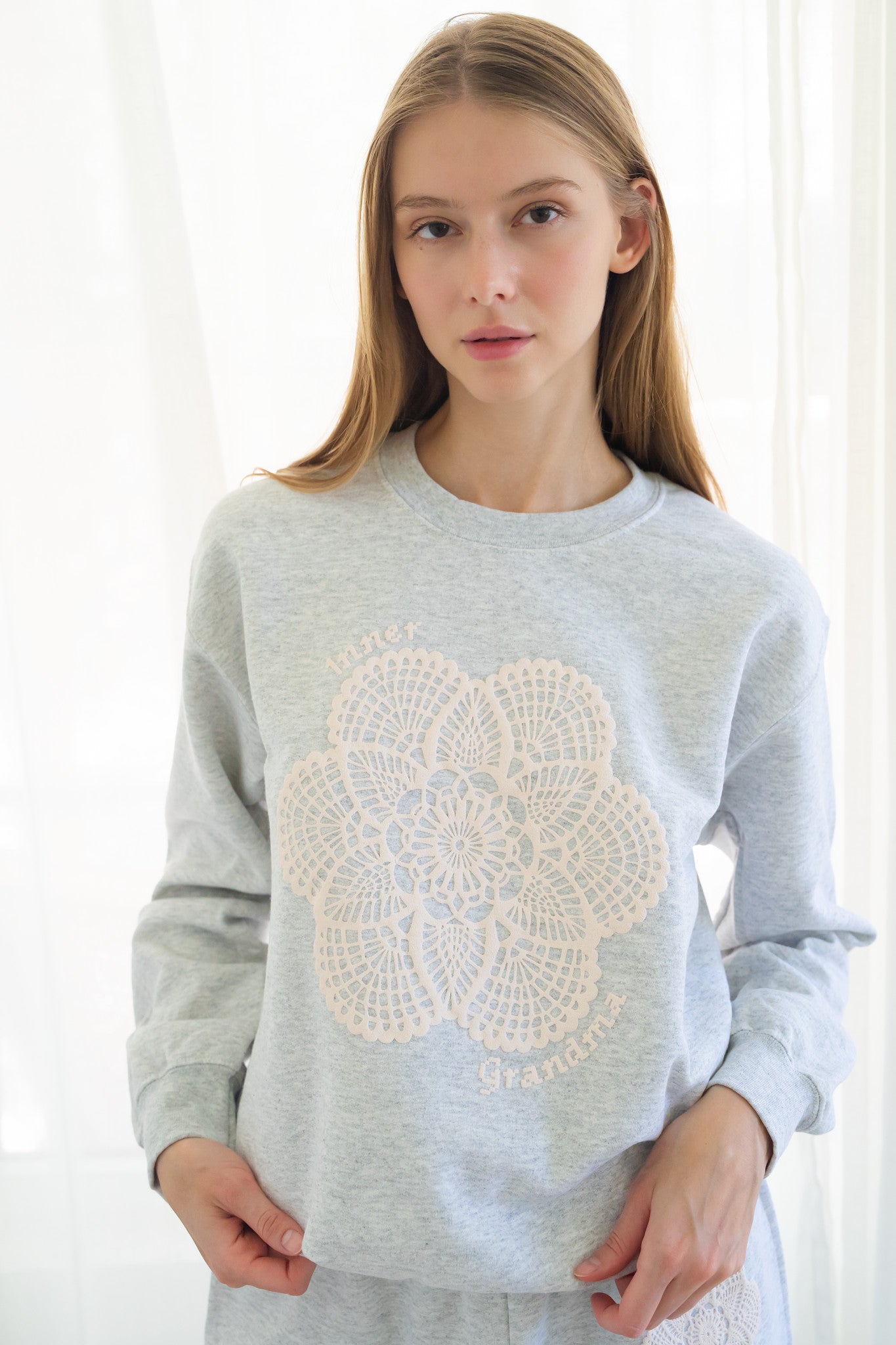 Person wearing a light gray crewneck sweatshirt with a large cream-colored crochet lace pattern on the front, set against a bright, softly lit background of white curtains.