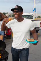 Person wearing a black hat with an embroidered emblem saying "Tired" holding a hot dog and a plate, standing outdoors near a grill and vehicles