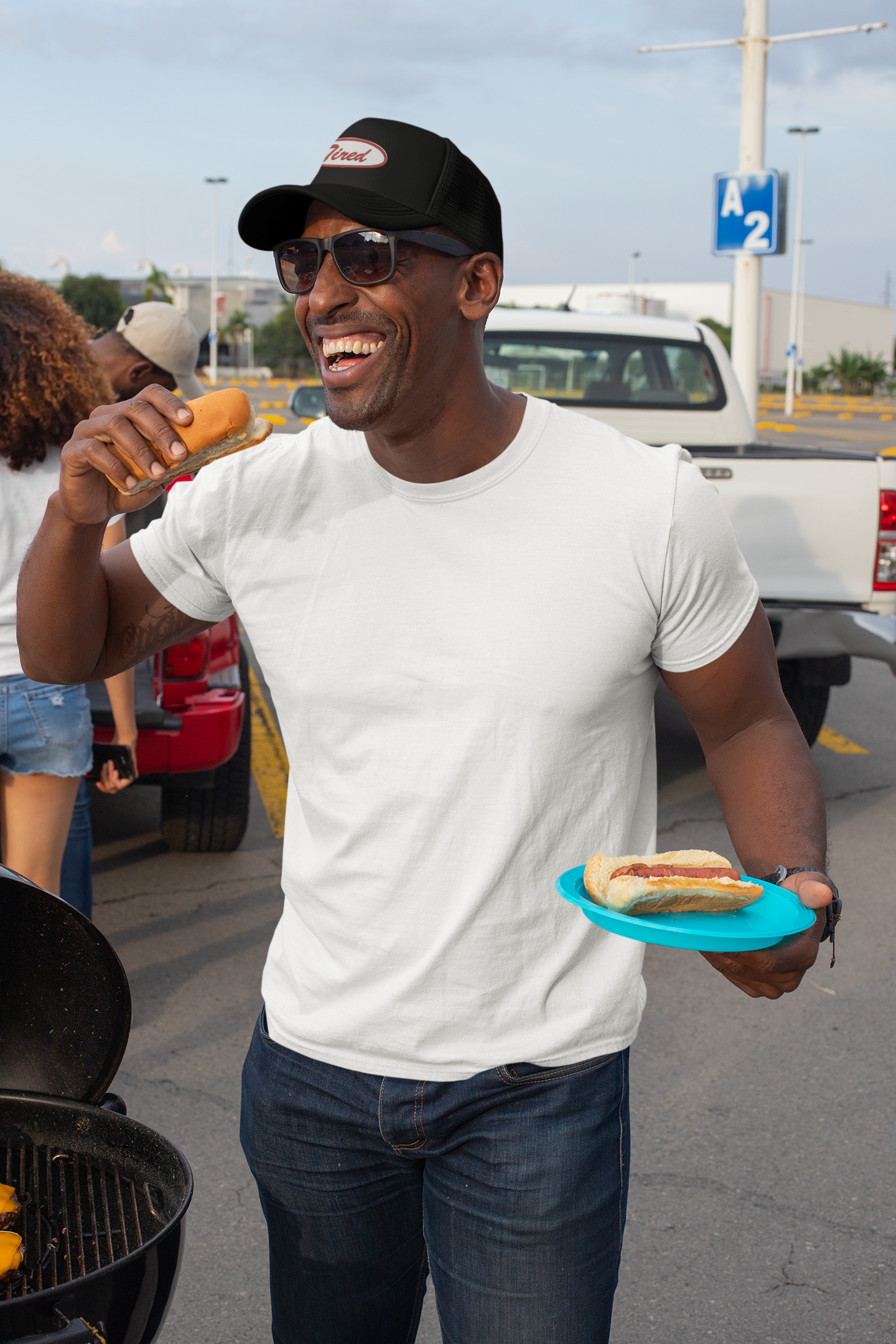 Person wearing a black hat with an embroidered emblem saying "Tired" holding a hot dog and a plate, standing outdoors near a grill and vehicles