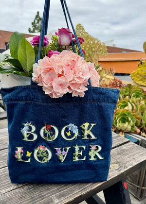 Blue denim tote bag with 'BOOK LOVER' text and floral design, held with flowers, on a wooden surface outdoors.