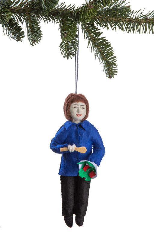 Ornament of Ina Garten in a blue coat holding a spoon and a bowl, hanging from a Christmas tree.