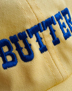 Close-up of a yellow cap with 'BUTTER' embroidered in blue thread.