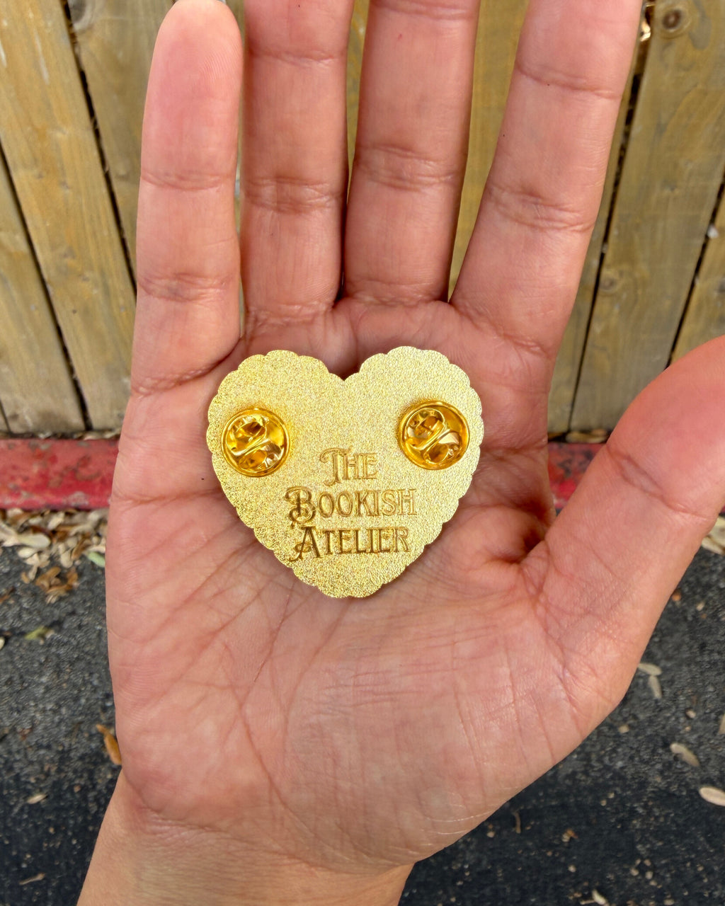 Heart-shaped gold pin with 'The Bookish Atelier' branding held in a hand against a wooden fence background.
