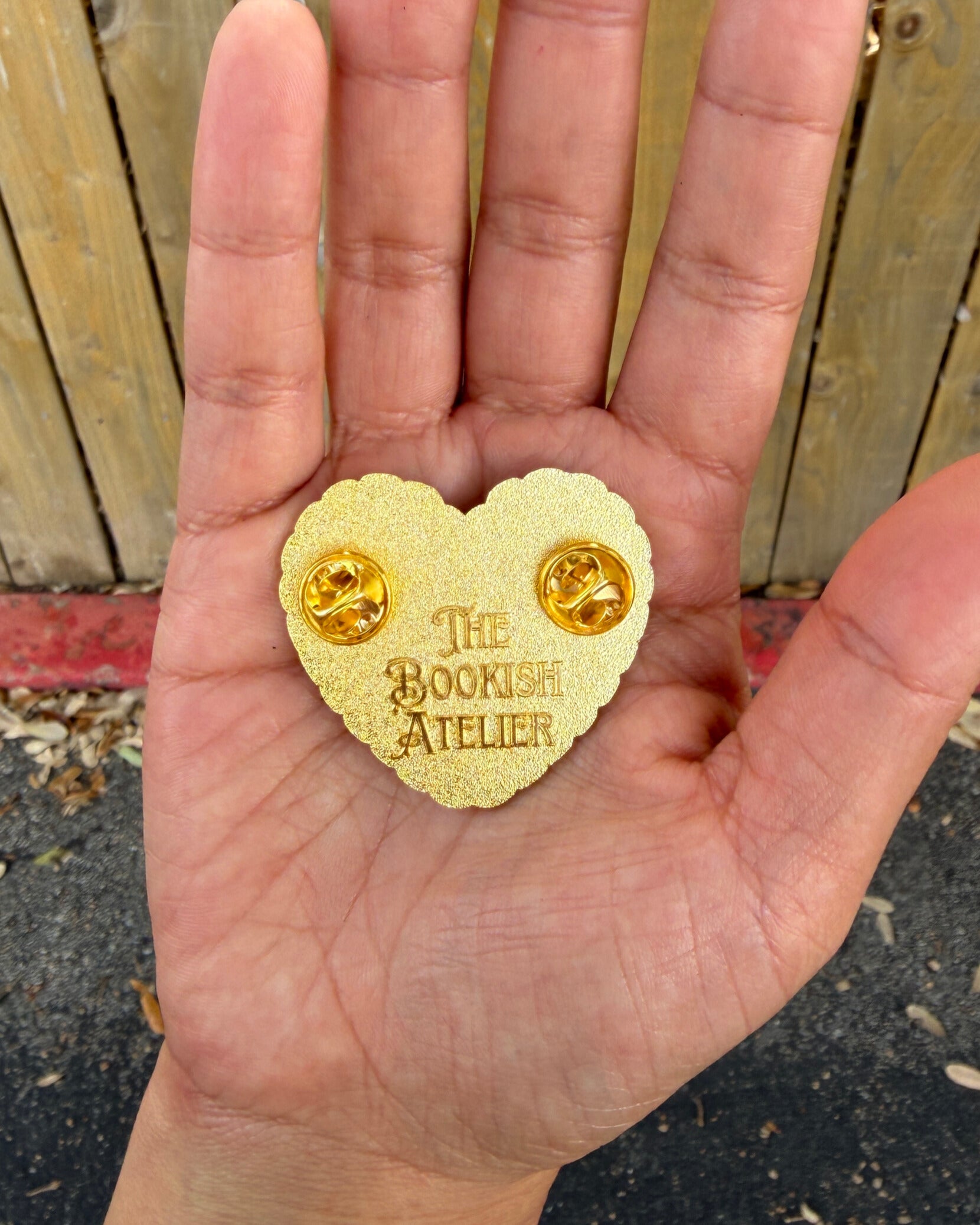 Heart-shaped gold pin with 'The Bookish Atelier' branding held in a hand against a wooden fence background.