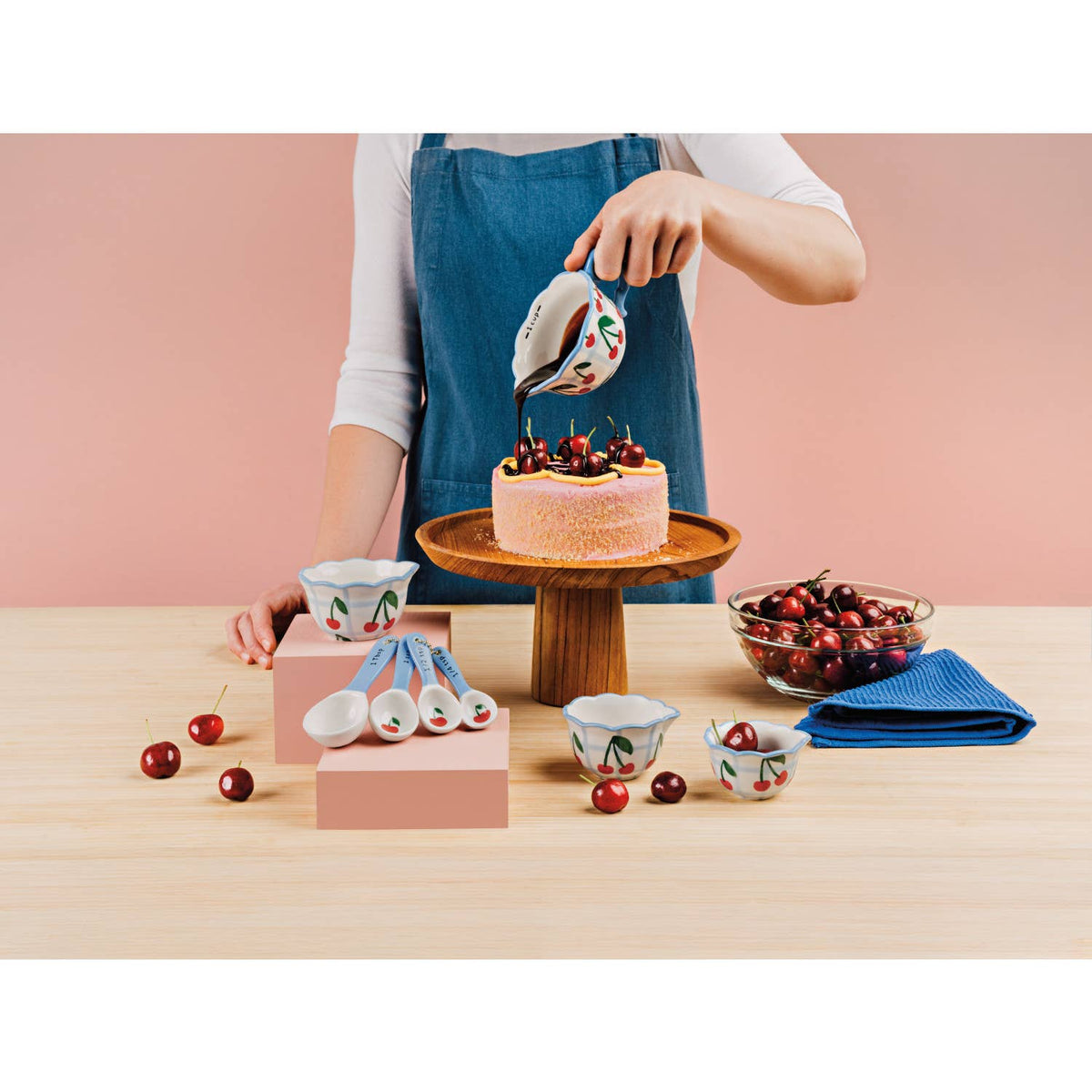 Person decorating a cake with cherries on a wooden table, surrounded by cherry-themed kitchenware.