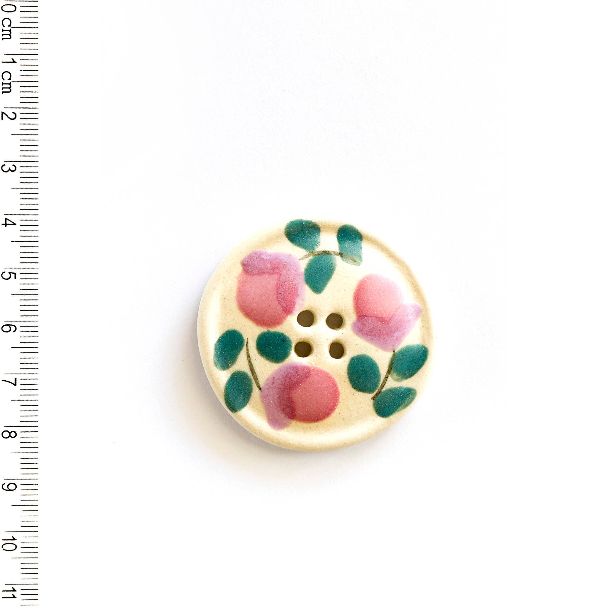 Off-white floral button with pink flowers and green leaves on a white background, next to a ruler for scale.