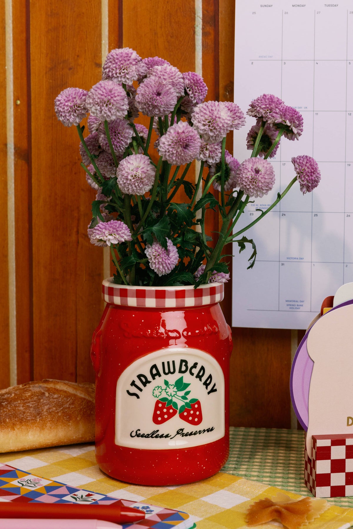 Red jar with strawberry preserve label filled with flowers on a table with bread