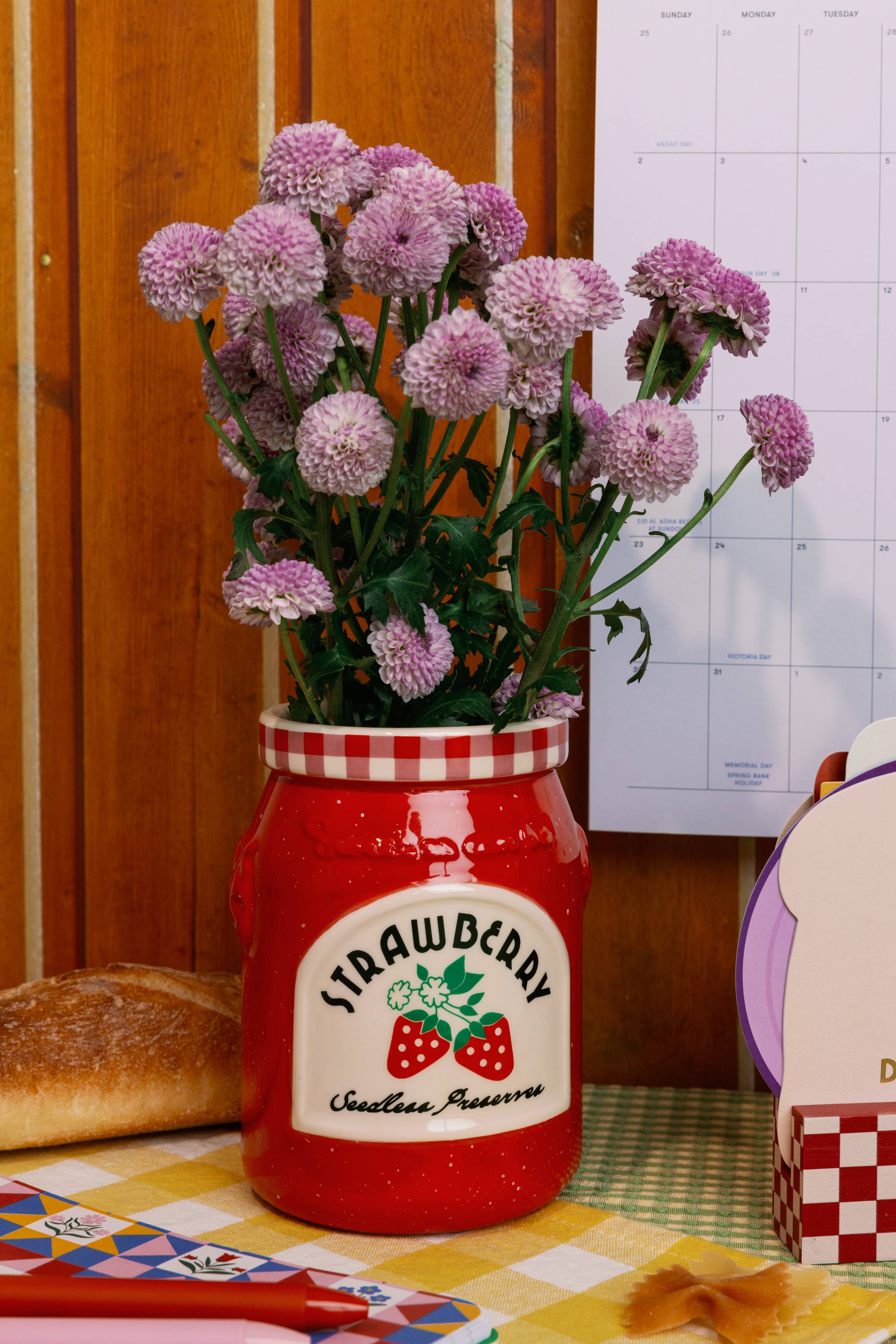 Red jar with strawberry preserve label filled with flowers on a table with bread