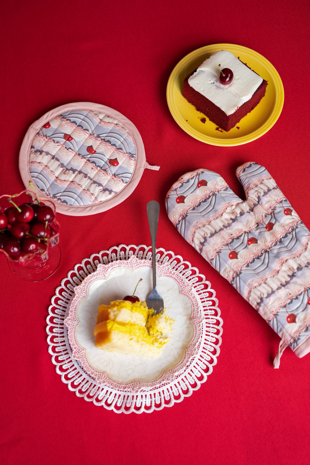 Dessert scene with a plate of cake, a bowl of cherries, and frosting printed oven mitts on a red background