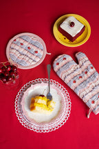 Dessert scene with a plate of cake, a bowl of cherries, and frosting printed oven mitts on a red background