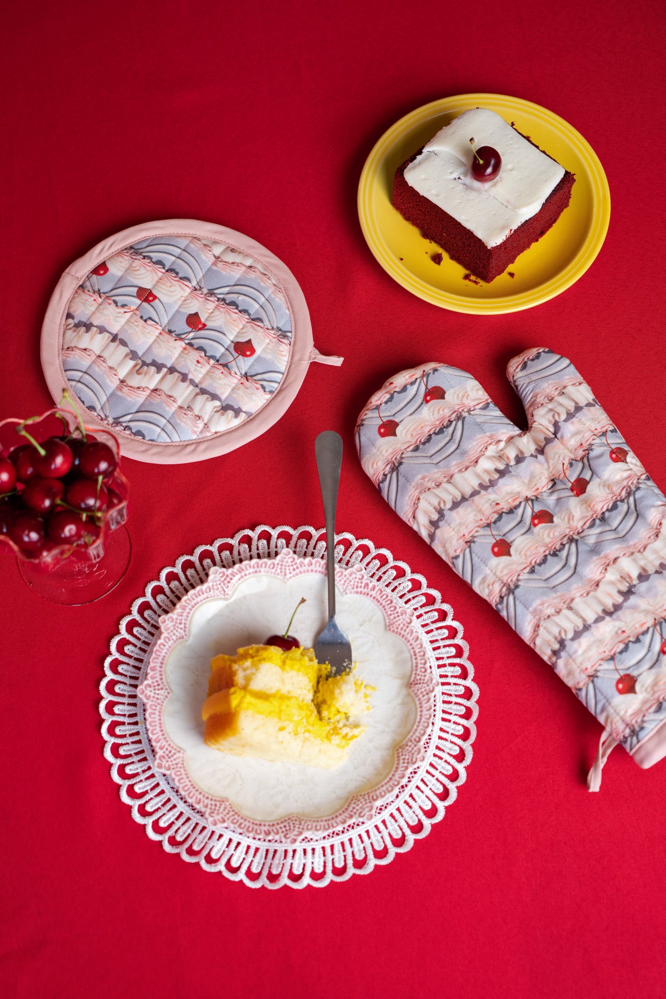 Dessert scene with a plate of cake, a bowl of cherries, and frosting printed oven mitts on a red background
