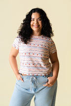 Person wearing a pink and purple frosting patterned shirt with cherries on it and light blue jeans against a beige background