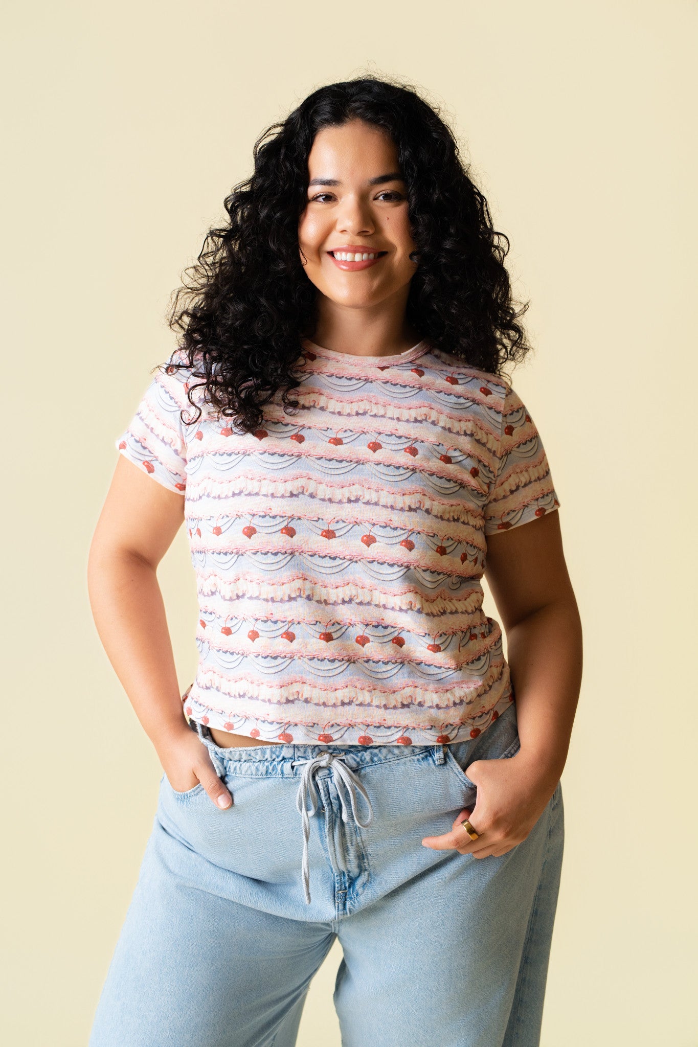 Person wearing a pink and purple frosting patterned shirt with cherries on it and light blue jeans against a beige background