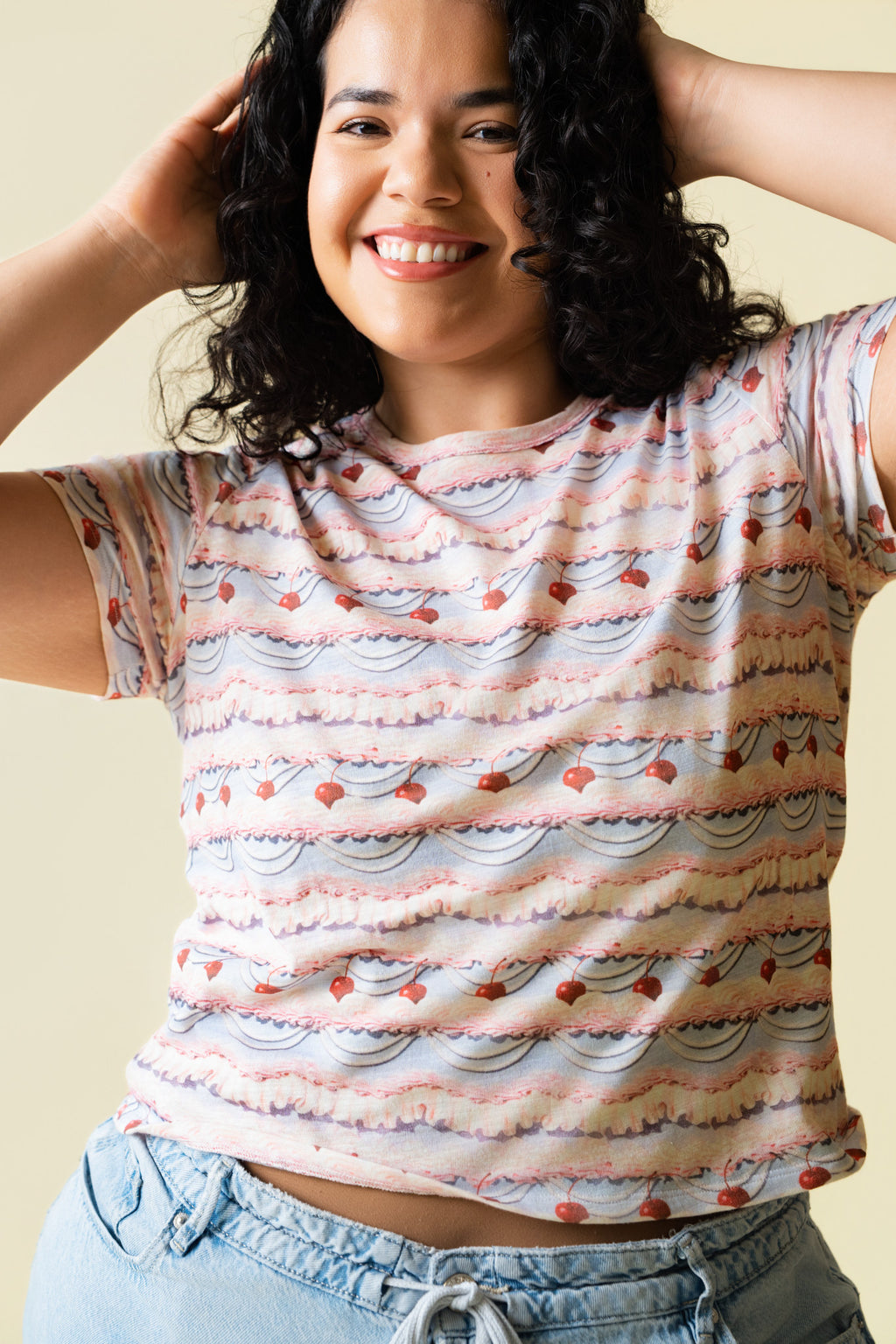 Person wearing a pink and purple frosting patterned shirt with cherries on it and light blue jeans against a beige 