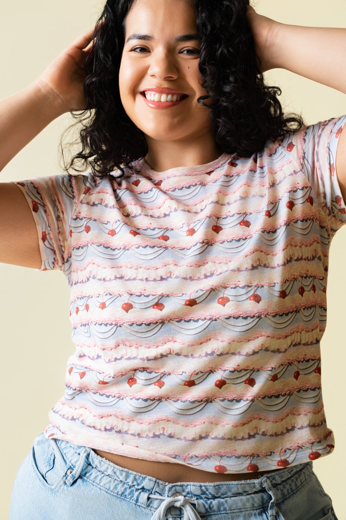 Person wearing a pink and purple frosting patterned shirt with cherries on it and light blue jeans against a beige 