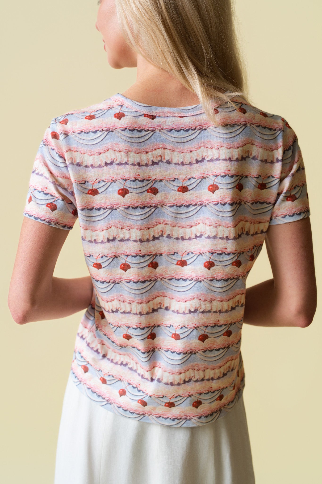 Back of a person wearing a pink and purple frosting patterned shirt with cherries on it and a white skirt on a beige back ground