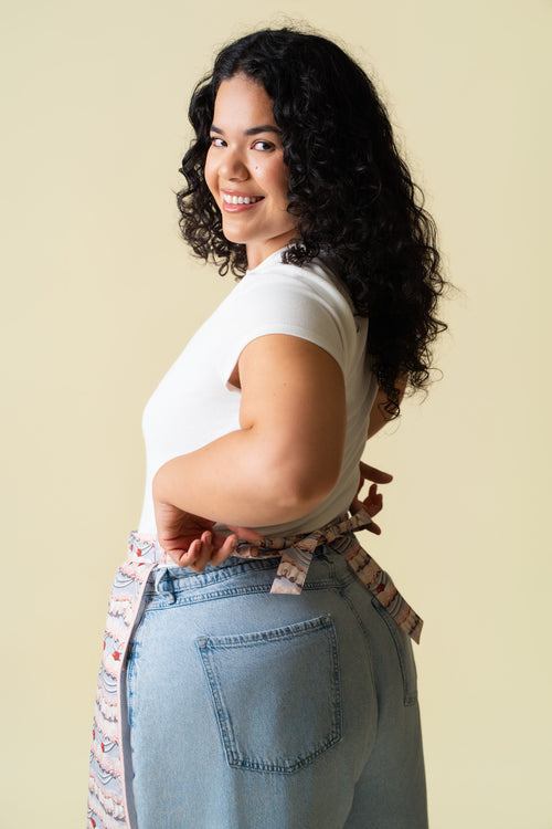 Person wearing a white t-shirt and light blue jeans with a frosting patterned apron against a beige background