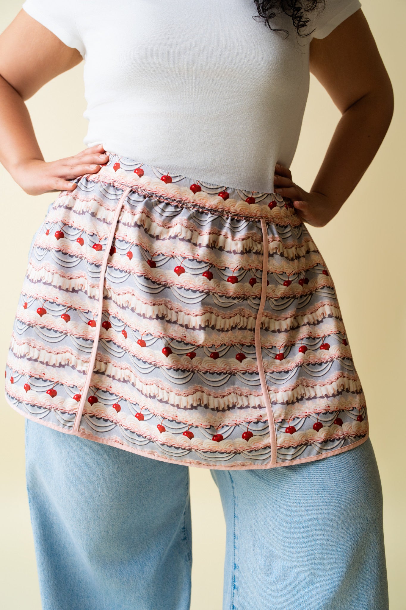 Person wearing a frosting patterned apron over a white shirt and blue jeans on a beige background.