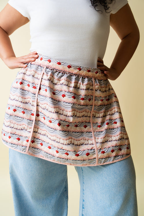 Person wearing a frosting patterned apron over a white shirt and blue jeans on a beige background.