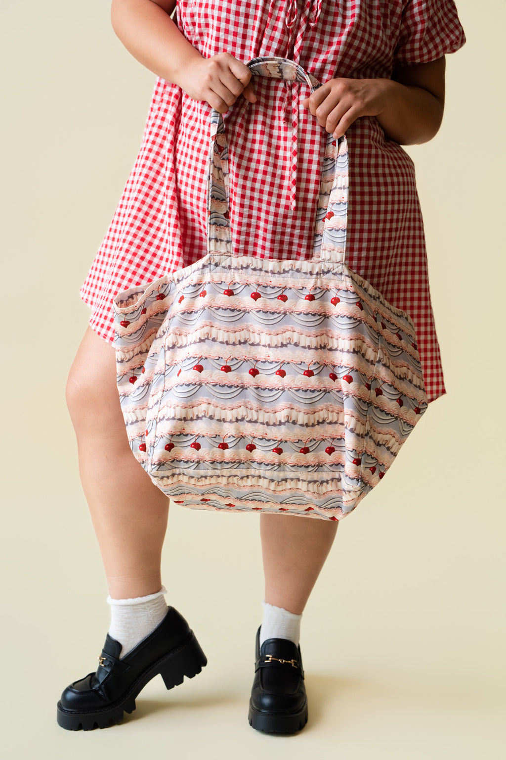 Person wearing a red checkered dress holding a tote bag with patterned fabric of decorative frosting with cherries on a beige background