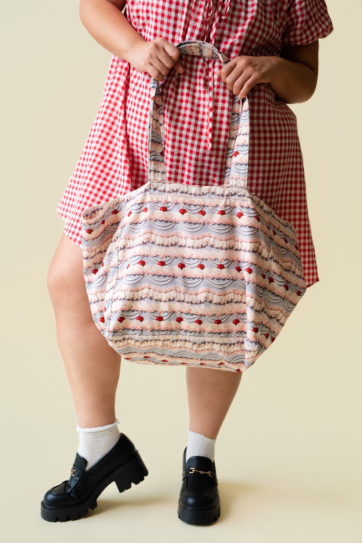 Person wearing a red checkered dress holding a tote bag with patterned fabric of decorative frosting with cherries on a beige background