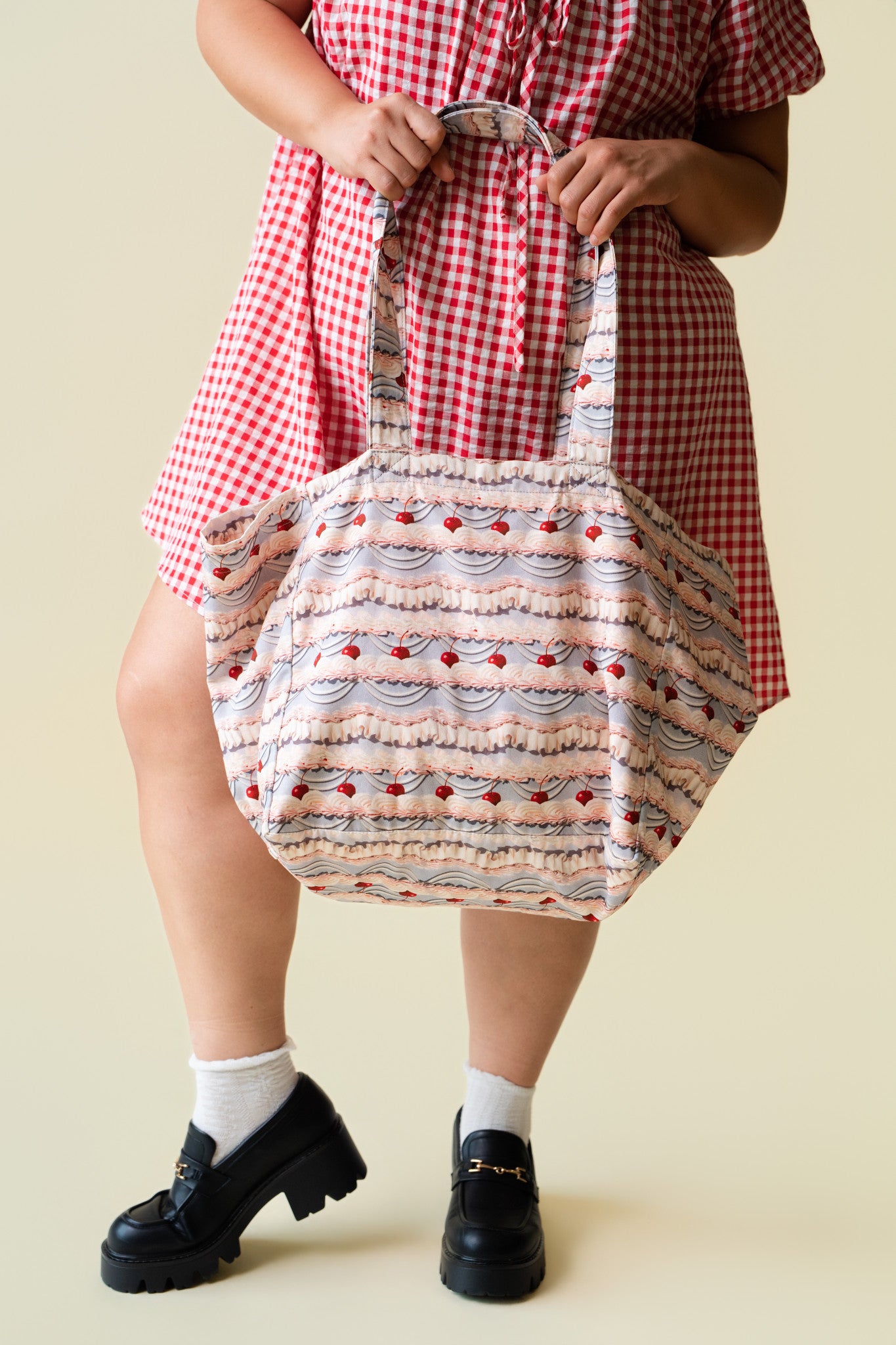 Person wearing a red checkered dress holding a tote bag with patterned fabric of decorative frosting with cherries on a beige background