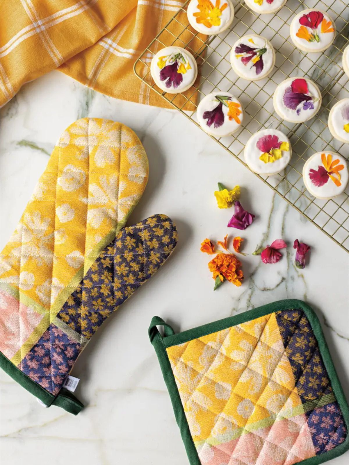 Quilted oven mitts and pot holder with floral patterns on a marble surface with decorated cupcakes.