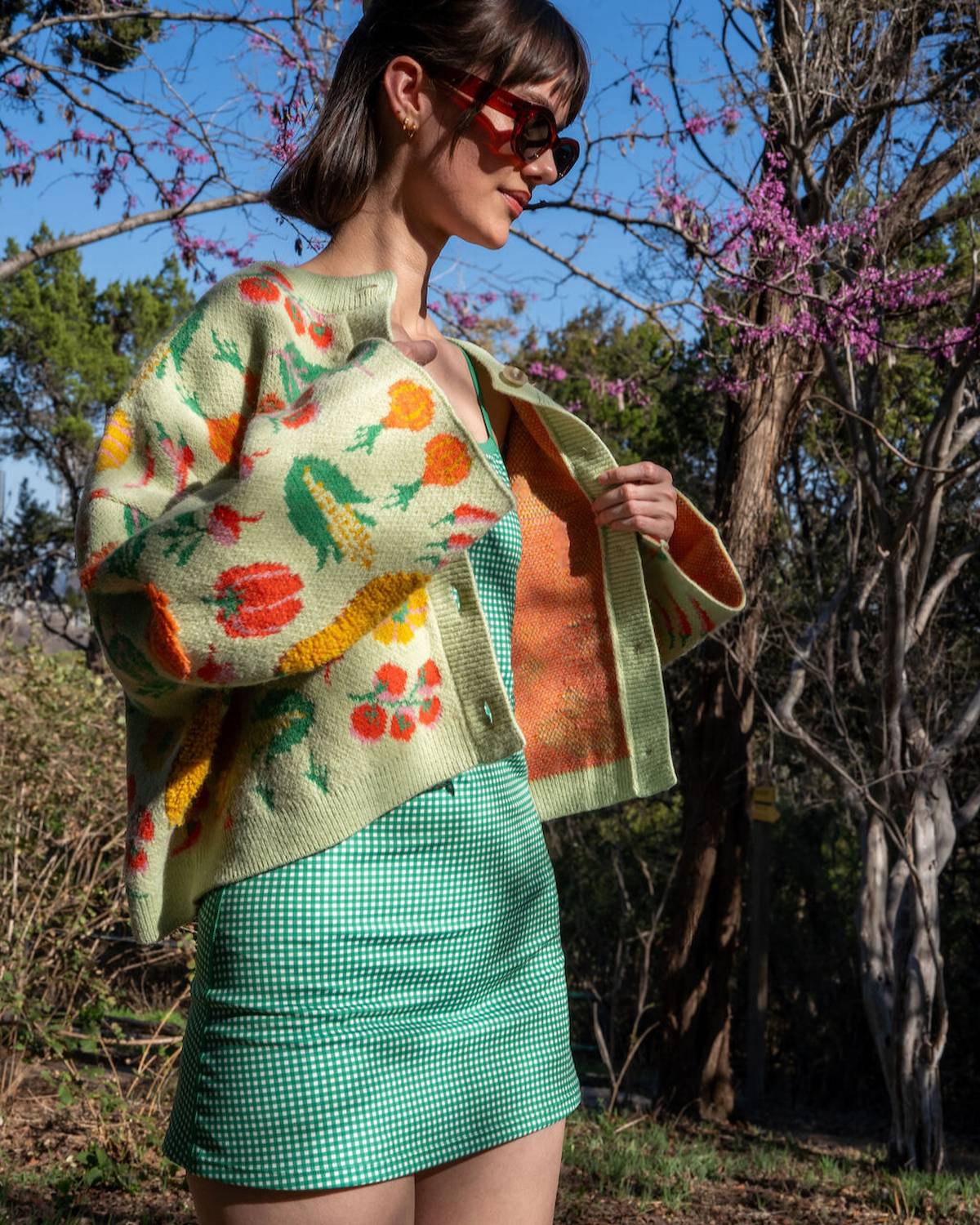 Woman wearing a colorful floral-patterned cardigan in a natural setting with trees and blue sky.